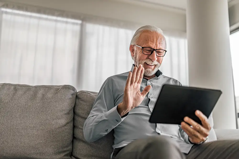 Cheerful senior man having video call on tablet sitting on the couch at home elderly man wearing eyeglasses staying in touch with friends and family using online video call connecting with people